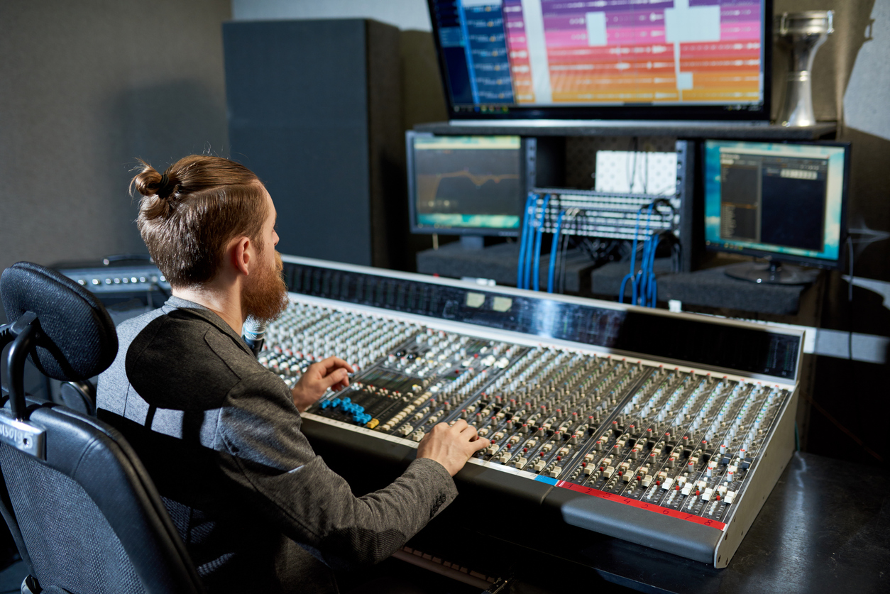 Un homme procède à divers réglages sur une console de mixage sonore. Au-dessus de celle-ci se trouvent plusieurs écrans d’ordinateur.