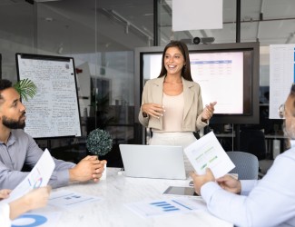 Image d'une alternante en entreprise, qui présente face à son équipe au travail des chiffres devant un écran et un paperboard.