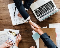 deux personnes se serrant la main, vue du dessus, il y a un ordinateur, des documents et une femme sur la gauche dont on ne voit que les mains