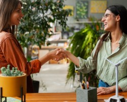 deux femmes qui se serrent la main dans un bureau, la femme de gauche porte un haut orange et celle de droite un haut vert. Elles se trouvent derrière un bureau, le meuble, dans un décor de bureau verdoyant.