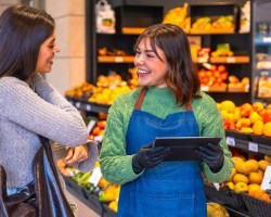une jeune femme avec un tablier discute avec une autre femme dans un supermarché, entourées de fruits et légumes