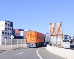 un camion orange sur une route, entrant dans ce qui ressemble à un port de commerce avec des conteneurs