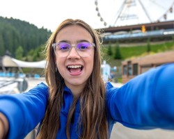une jeune fille portant des lunettes et un sweat bleu tenant une caméra que l'on ne voit pas (nous sommes la caméra). Elle se trouve dans un parc d'attractions.