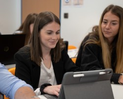 Deux jeunes femmes dans une salle de cours regardent une tablette, elles sont assisent à une table.